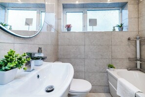 Ein modernes Badezimmer mit einem großen Waschbecken, einer Toilette und einer Badewanne. Zwei Fenster mit Blumentöpfen auf den Fensterbänken.