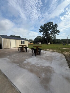 A concrete patio with a table and chairs, a building with a garage door, and a large tree in the background.