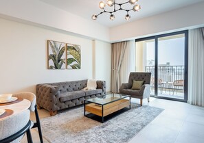 A modern living room with a grey sofa, armchair, and coffee table. There are two framed plant artworks on the wall, a large window with curtains, and a balcony visible through the window. - 1BR Umm Suqiem Apartment w/ Burj Al Arab Pool View, Steps to Kite Beach (Dubai)