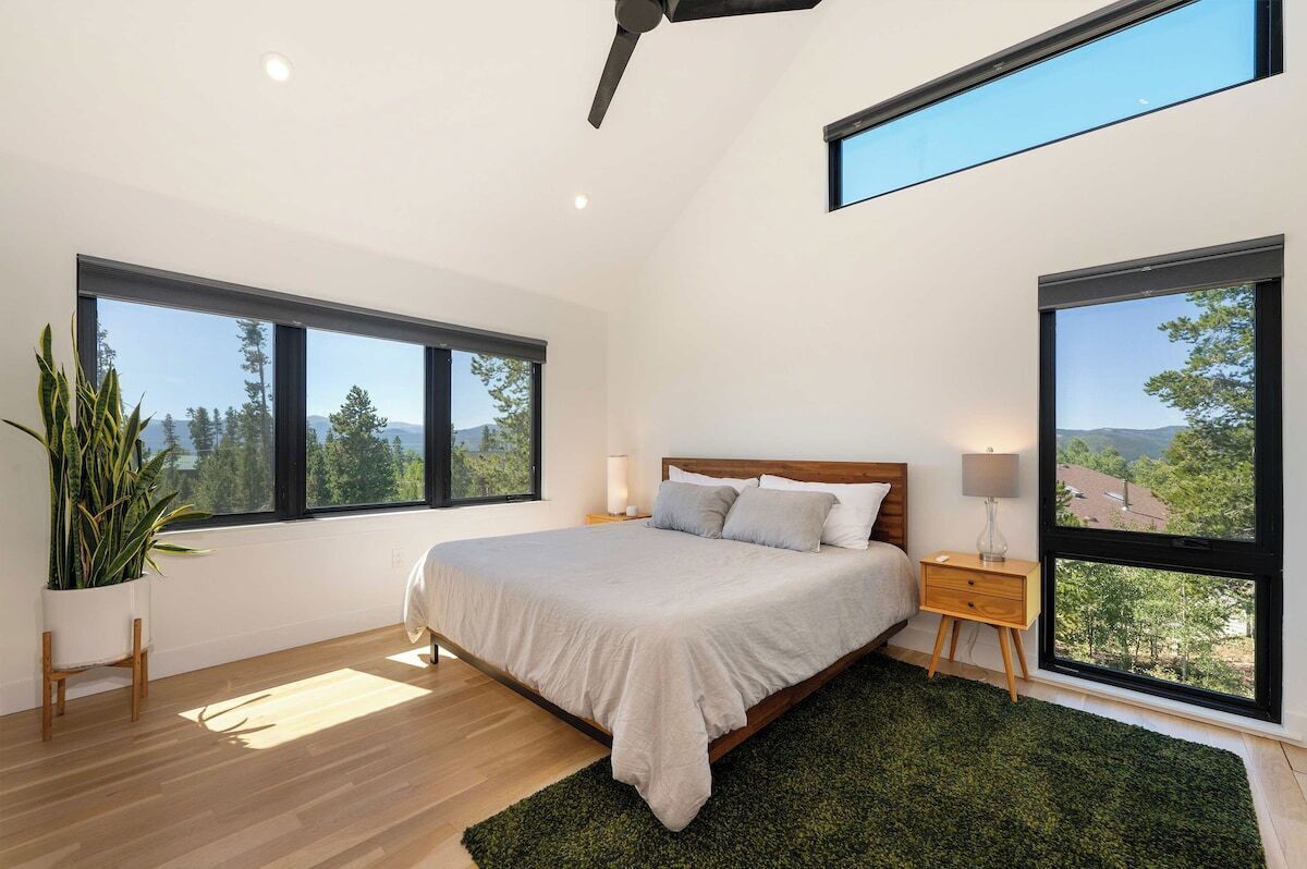 A bedroom with a large bed, two bedside tables, a ceiling fan, and large windows offering a view of trees and mountains.