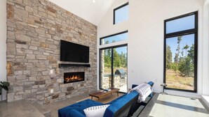 A modern living room with a stone fireplace, a flat-screen TV, a blue sofa, and large windows offering a view of the outdoors. - Ridgeline Retreat | Modern 4BR, Big Mountain Views (Tabernash)