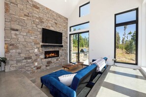 Ein modernes Wohnzimmer mit Kamin aus Stein, einem Flachbildfernseher, einem blauen Sofa und großen Fenstern mit Blick ins Freie.