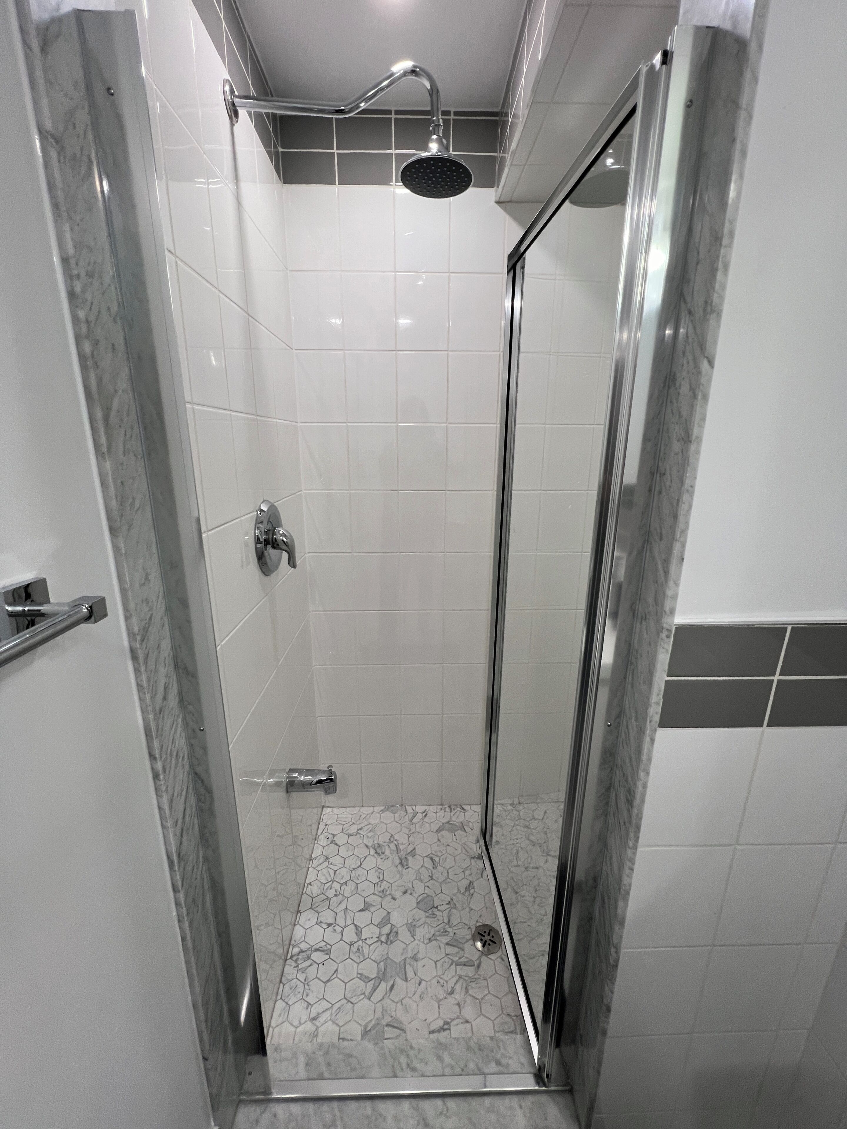 A glass-enclosed shower with a handheld showerhead, white tiled walls, and a marble floor.