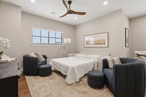 Ein Schlafzimmer mit einem Bett, zwei Sesseln, einem Nachttisch, einer Stehlampe, einem Deckenventilator, einem Fenster mit Jalousien und einem gerahmten Bild an der Wand.