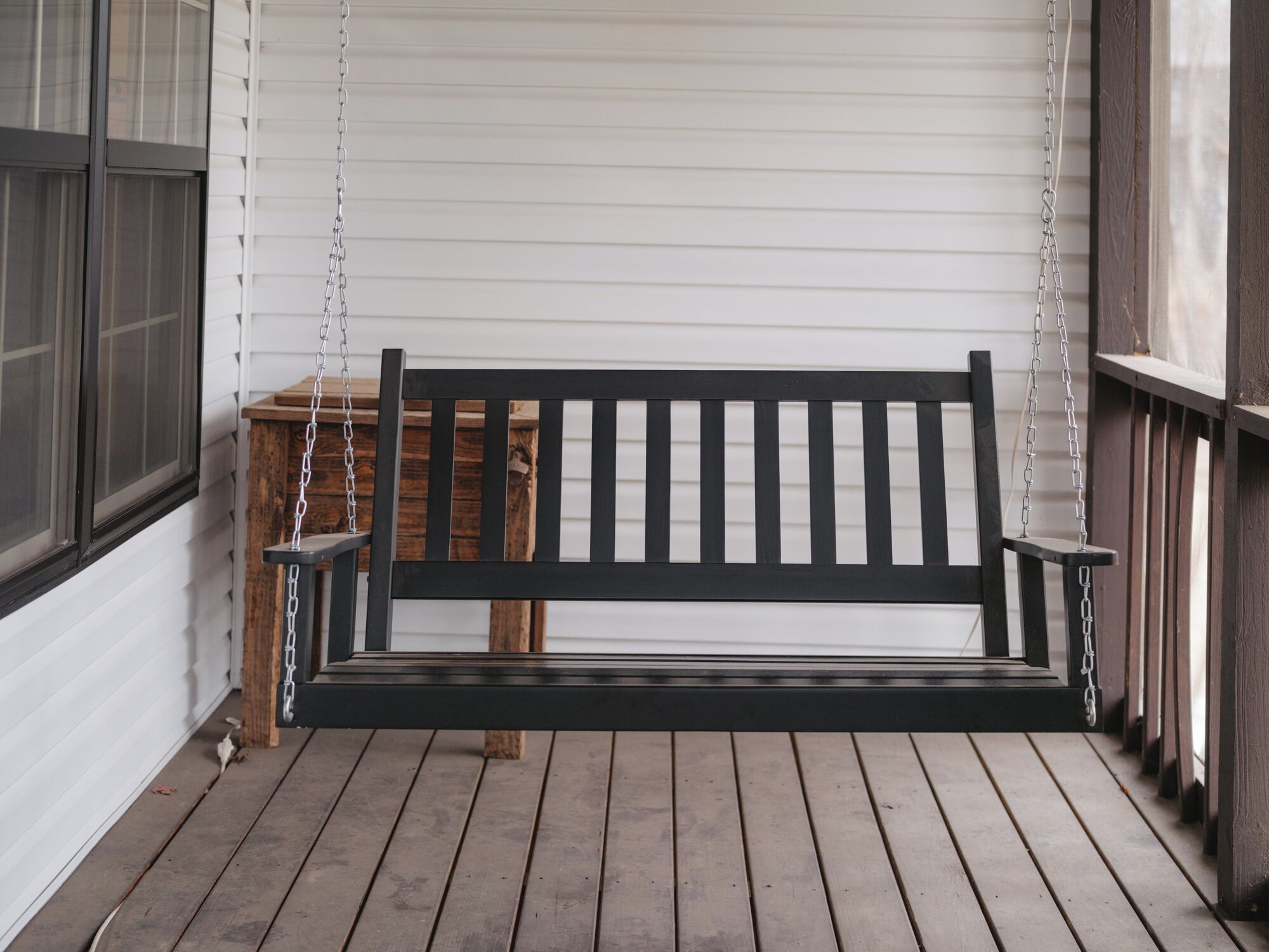 A wooden porch with a black bench, white siding, and a wooden table.