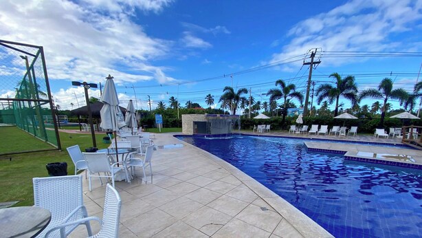 A swimming pool with tiled edges, surrounded by white wicker chairs and tables, palm trees, and a clear sky. - Muro Alto Condomínio Clube por YOLO (Ipojuca)