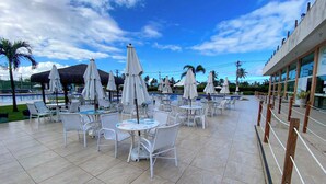 An outdoor pool area with white chairs and umbrellas, a tiled floor, and a building with large windows in the background. - Muro Alto Condomínio Clube por YOLO (Ipojuca)