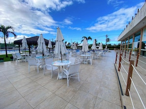 An outdoor pool area with white chairs and umbrellas, a tiled floor, and a building with large windows in the background. - Muro Alto Condomínio Clube por YOLO (Ipojuca)