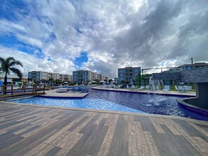 A swimming pool area with a wooden deck, lounge chairs, and a palm tree. - Muro Alto Condomínio Clube por YOLO (Ipojuca)