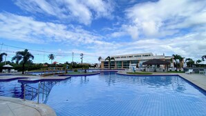 A swimming pool area with multiple sections, a covered seating area, and a building in the background. - Muro Alto Condomínio Clube por YOLO (Ipojuca)