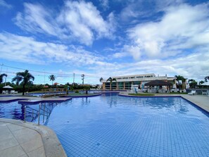 A swimming pool area with multiple sections, a covered seating area, and a building in the background. - Muro Alto Condomínio Clube por YOLO (Ipojuca)
