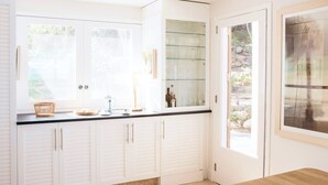 Una cocina con muebles blancos, un armario con puerta de vidrio que contiene copas de vino y una ventana con vistas a la vegetación.