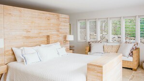 Ein Hotelzimmer mit einem großen Bett, einem Holzkopfende, einem Nachttisch, einer Lampe, einem Fenster mit Jalousien und einem Korbstuhl.