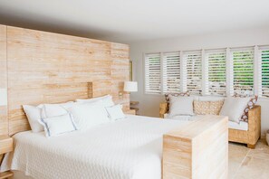 A hotel room with a large bed, a wooden headboard, a nightstand, a bedside table, a lamp, a window with blinds, and a wicker chair. - Villa Marie (2 bedrooms) (Saint Barthélemy)