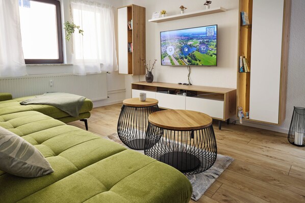 A modern living room with a large flat-screen TV, green sofas, a wooden coffee table, and a decorative plant.