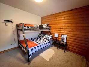 A bunk bed with a checkered bedspread, a small bedside table with a lamp, and a wall-mounted shelf.