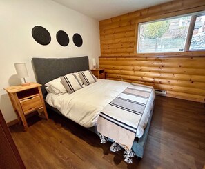 A bedroom with a wooden log wall, a bed with a striped blanket, a nightstand with a lamp, and a window with blinds. - Lakeview Retreat with Buoy (Celista)