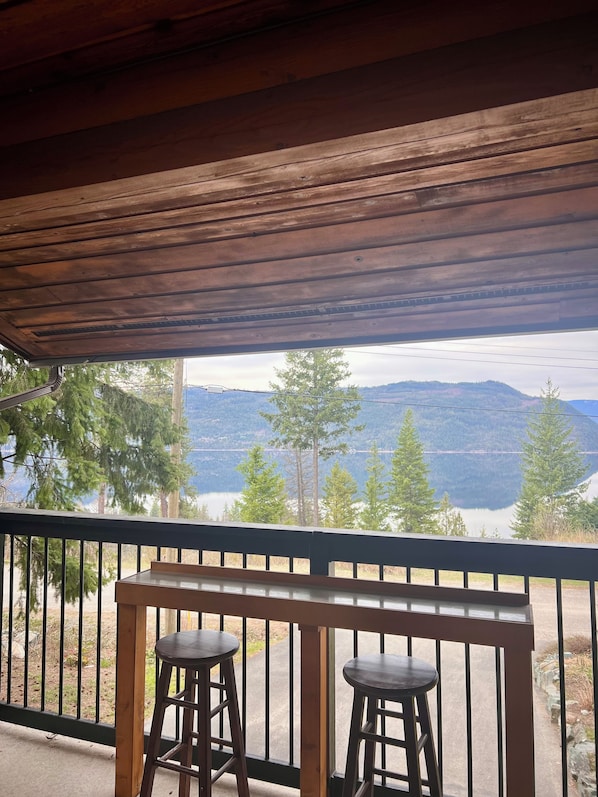 A wooden balcony with two stools, overlooking a lake and mountains.