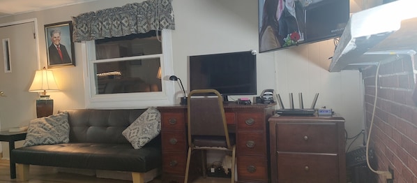 A living room with a sofa, a desk with a computer and a printer, a TV mounted on the wall, and a window with curtains.