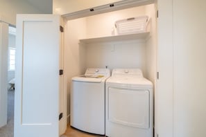 A compact laundry room with two white front-loading washing machines and a white laundry basket on a shelf. - Seascape Haven, Modern Townhome with Fireplace (Depoe Bay)