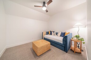 Une pièce avec un ventilateur de plafond, un canapé bleu avec des coussins, un pouf en osier, une petite table d’appoint avec une lampe et une plante en pot.