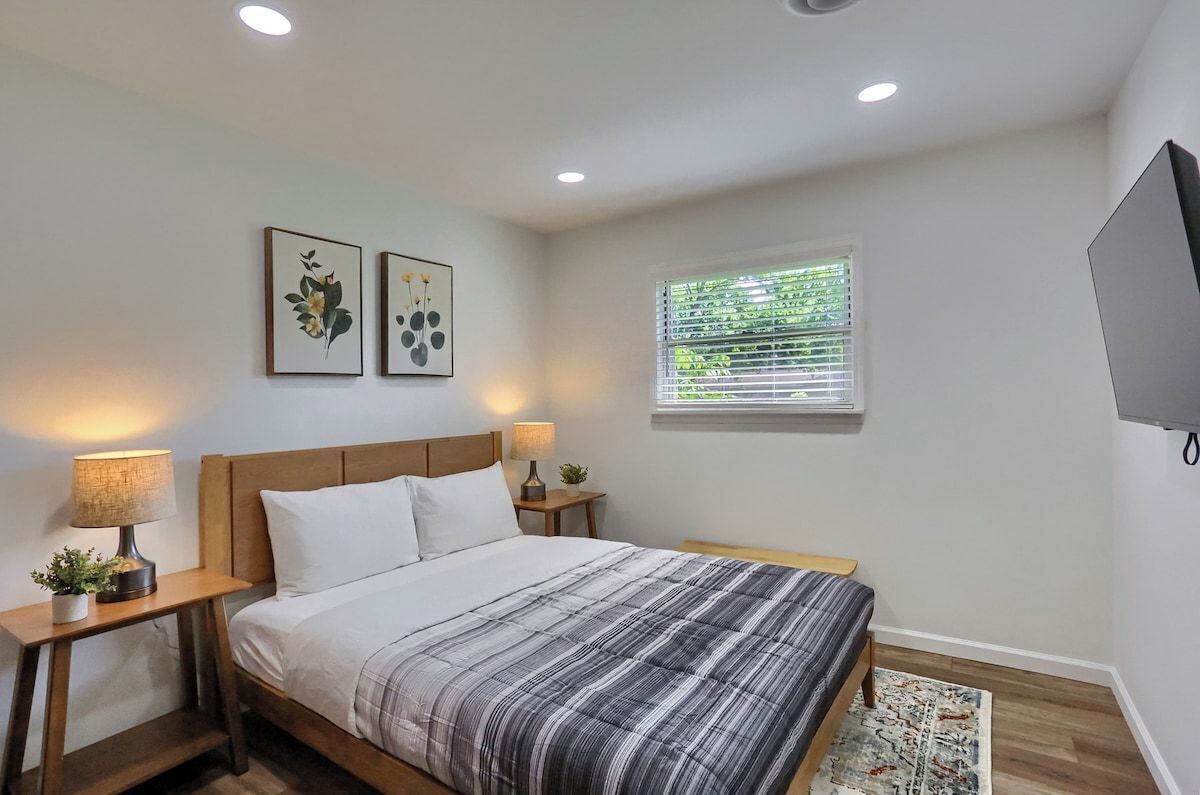 Un dormitorio con cama, dos mesitas de noche, televisor montado en la pared y ventana con persianas.