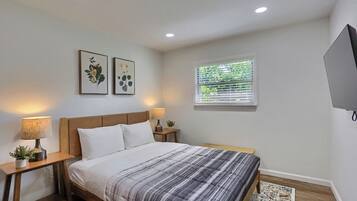 Un dormitorio con cama, dos mesitas de noche, televisor montado en la pared y ventana con persianas.