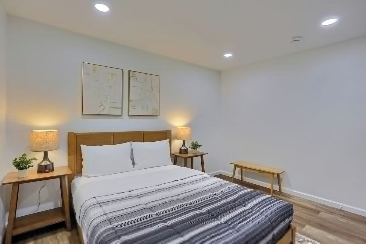 Une chambre à coucher comprenant un lit, deux tables de chevet avec des lampes, un banc et deux œuvres d’art accrochées au mur.