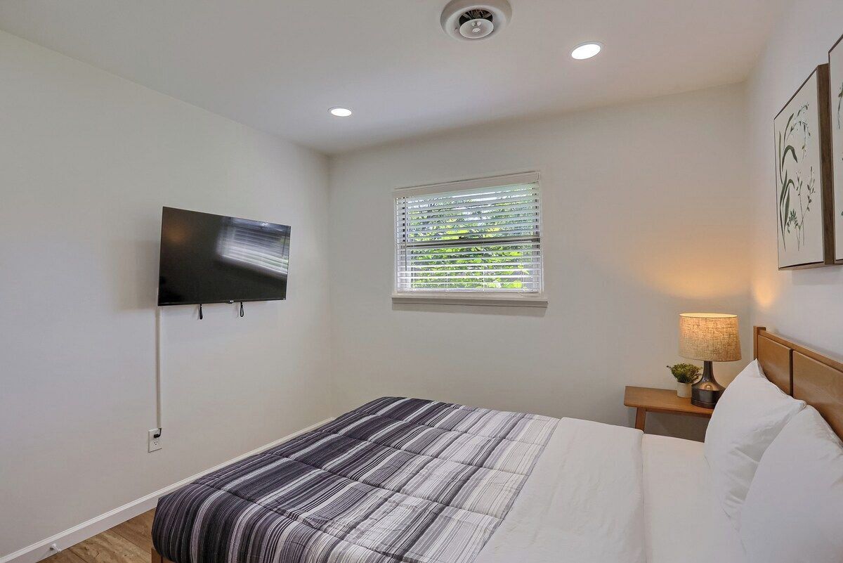 Une chambre à coucher avec un lit, un téléviseur fixé au mur, une fenêtre avec des stores, une lampe de chevet et une petite table sur laquelle est posée une plante.