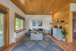 A modern interior with a wooden ceiling, a grey sofa, a kitchen area, and a large window with a view of trees. - Beautiful Dog Friendly Cabin in Fairplay (Fairplay)