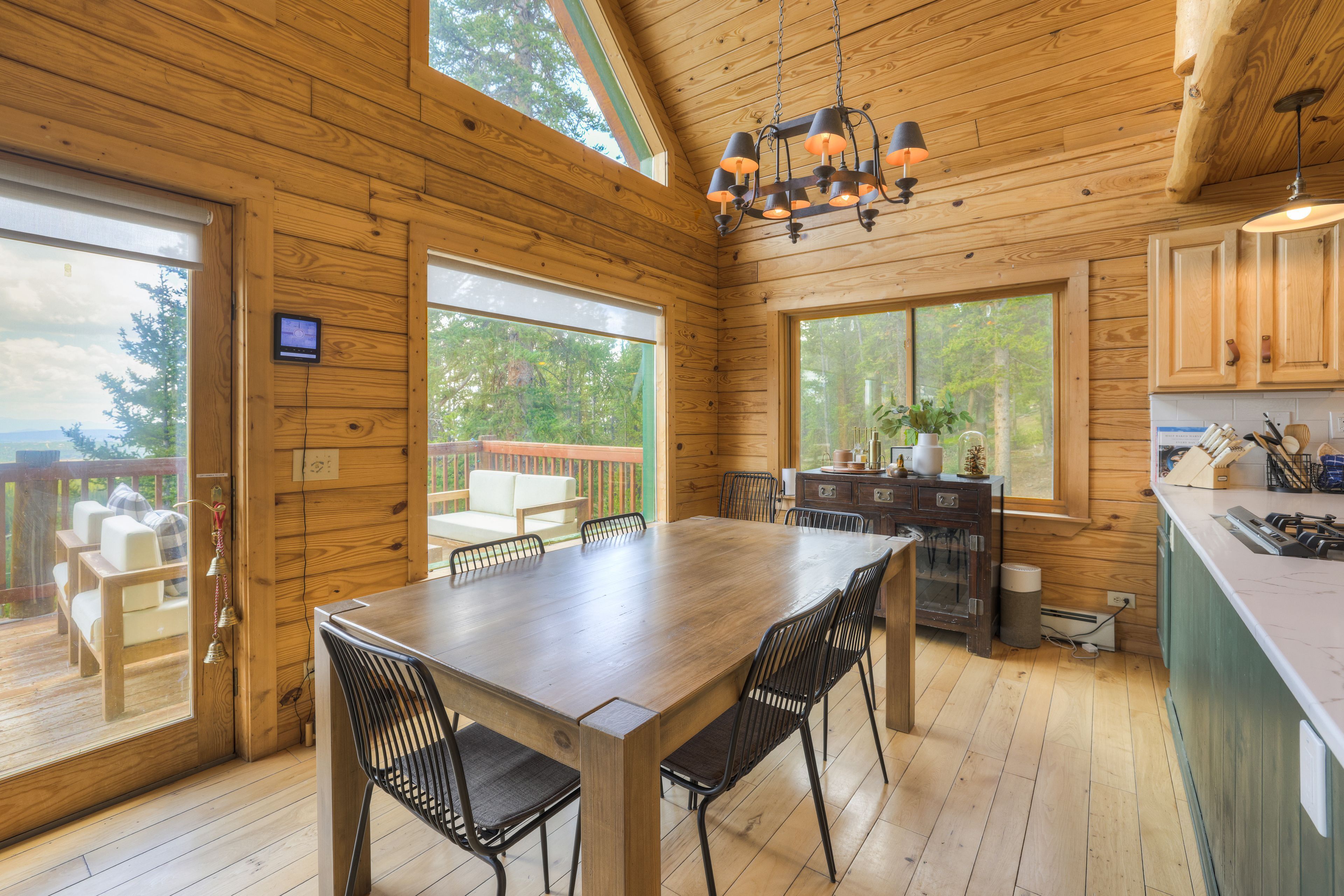 A wooden cabin interior with a dining table, chairs, a kitchen area, and large windows offering views of the outdoors.