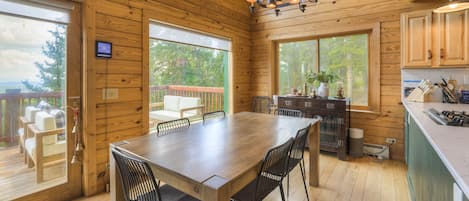 A wooden cabin interior with a dining table, chairs, a kitchen area, and large windows offering views of the outdoors.