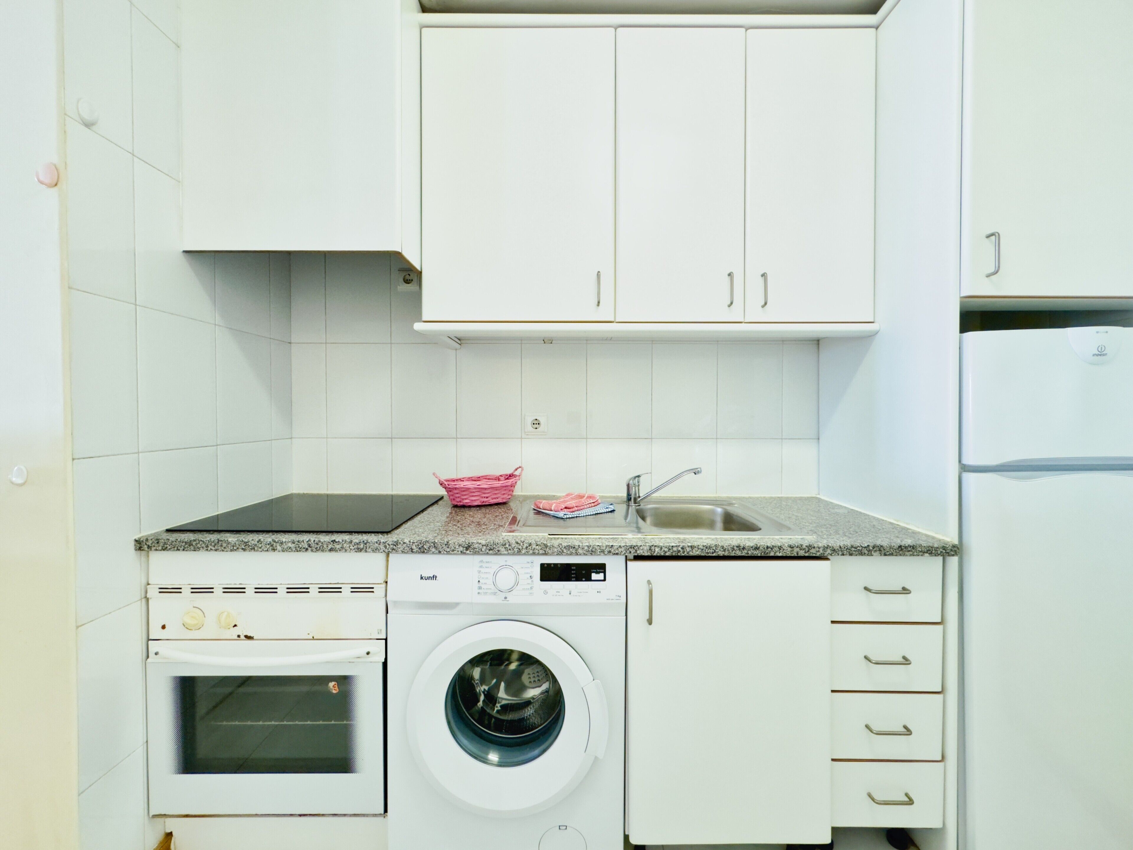 Una cocina compacta con armarios blancos, horno empotrado, lavadora y un lavaplatos.