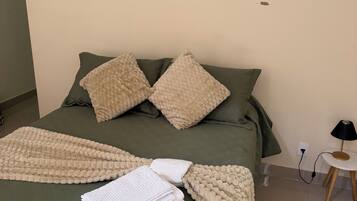 A neatly made bed with a green bedspread and two beige pillows, a folded white towel, and a blanket on the bed.