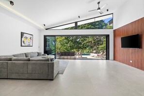 Un salon moderne avec un canapé d’angle gris, un téléviseur à écran plat fixé à un mur en bois, et une grande porte vitrée donnant sur une terrasse extérieure avec une piscine et des chaises longues.
