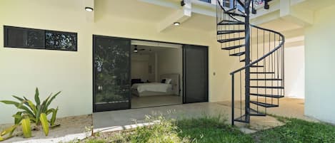 Una casa moderna con una escalera espiral, un dormitorio con cama y mesita de noche, y un pequeño jardín con plantas.