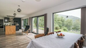 A modern dining area with a table set for four, a kitchen in the background, and a large window with a view of greenery and mountains. - Taigh Creag Thulach (Taynuilt)