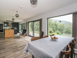 A modern dining area with a table set for four, a kitchen in the background, and a large window with a view of greenery and mountains. - Taigh Creag Thulach (Taynuilt)