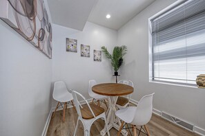 Een eetruimte met een ronde houten tafel en witte stoelen, een potplant en wanddecoratie.