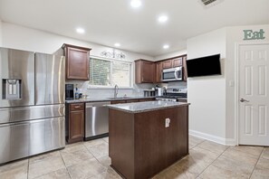 A modern kitchen with stainless steel appliances, wooden cabinets, a granite countertop island, and a flat-screen TV mounted on the wall.