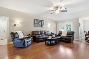 A living room with a brown leather sofa, a recliner, and a coffee table. There is a ceiling fan, wall-mounted lights, and a door.