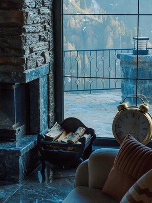 Una habitación con chimenea de piedra, ventana con vistas a un paisaje montañoso, barandilla metálica en el exterior y un reloj despertador antiguo.