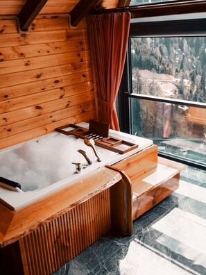 Interior de una cabaña de madera con una bañera grande, paredes de madera y una ventana con vistas a un bosque.