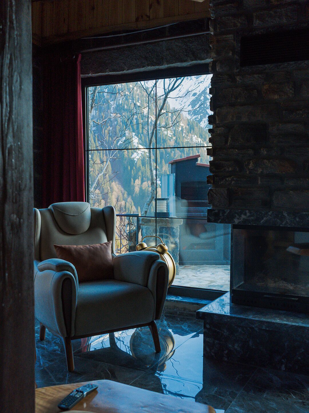Une pièce chaleureuse avec une cheminée en pierre, un fauteuil confortable et une vue sur un paysage enneigé par une grande fenêtre.