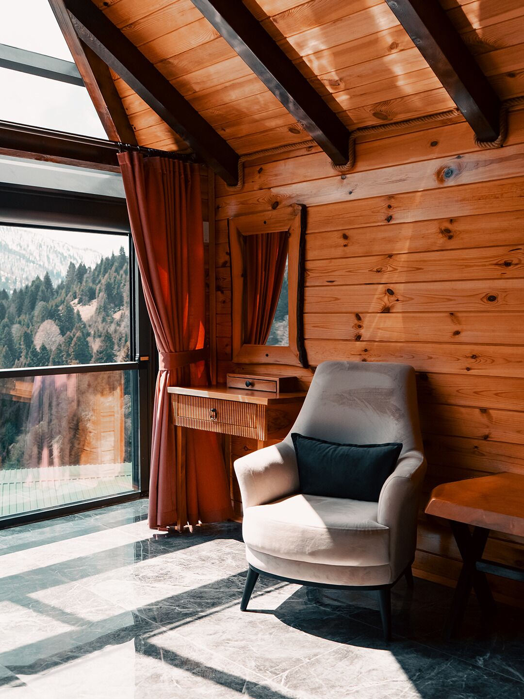 Une pièce avec un plafond en bois, une grande fenêtre avec des rideaux, une chaise avec un coussin et une table.