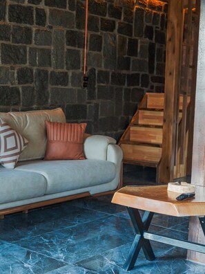 Una habitación con pared de piedra, un sofá con cojines, una mesa de madera y un control remoto, además de una escalera.