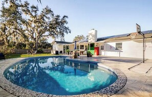 A swimming pool with a stone border, a house with a green door, and a basketball hoop.
