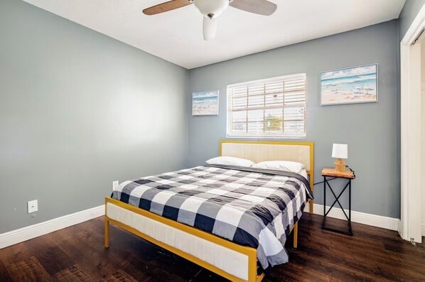 A bedroom with a bed, a ceiling fan, a window with blinds, and two framed pictures on the wall.