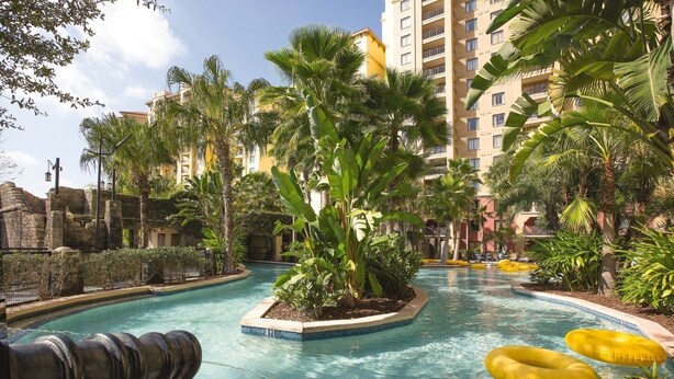 A pool area with palm trees, a stone wall, and a building in the background. - Orlando | Pool & Disney Access | 2BR Suite (Lake Buena Vista)