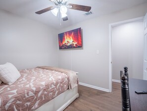 A bedroom with a bed, a ceiling fan, a painting of a fire, and a door.